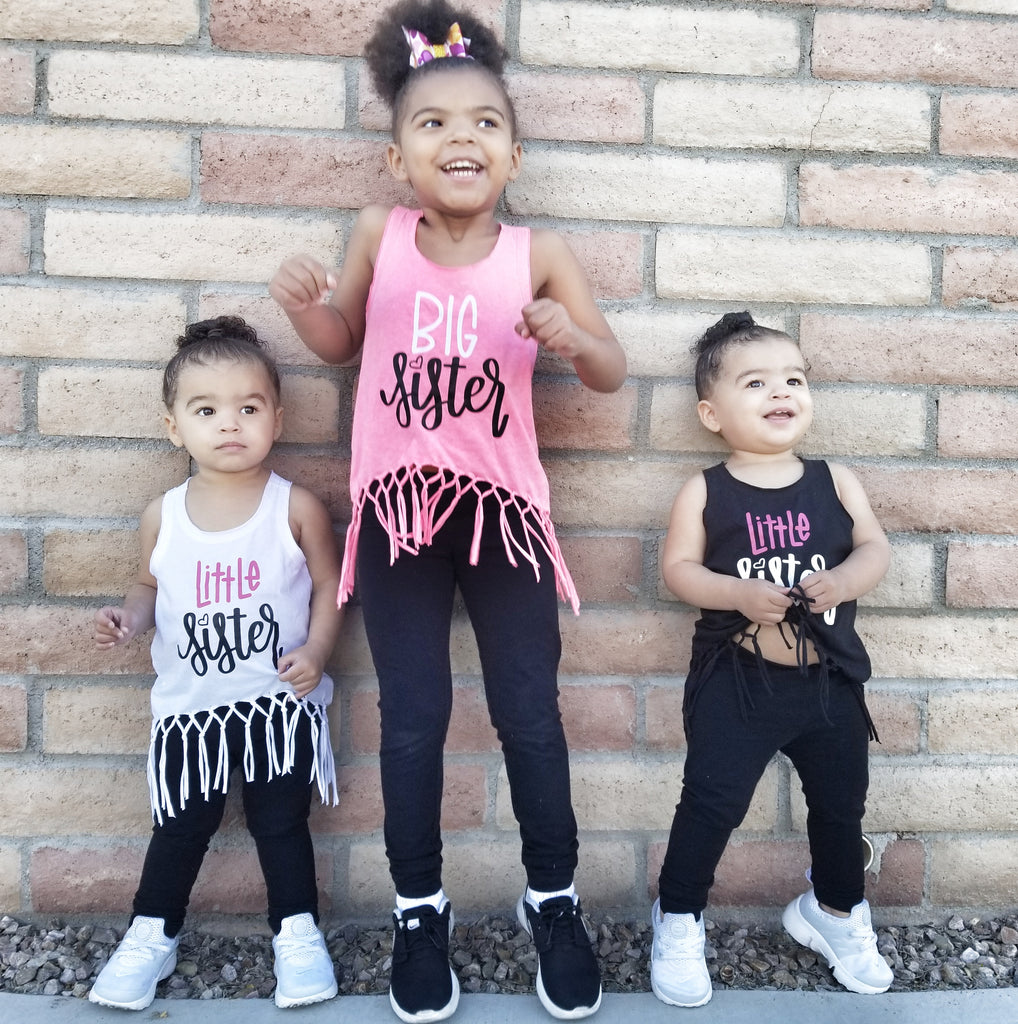 Little girl wearing pink fringe shirt that says Big Sister in white and black with twin toddler girls wearing white and black fringe tanks that say Little Sister in white and pink