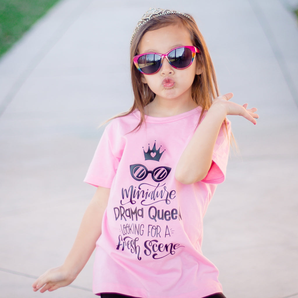 Girl wearing sunglasses, tiara, and a shirt that says miniature drama queen in black