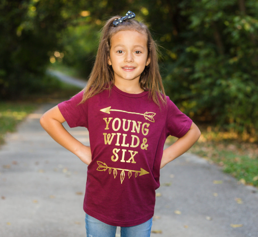 Little girl wearing wine colored shirt with Young Wild and Six in gold with arrows