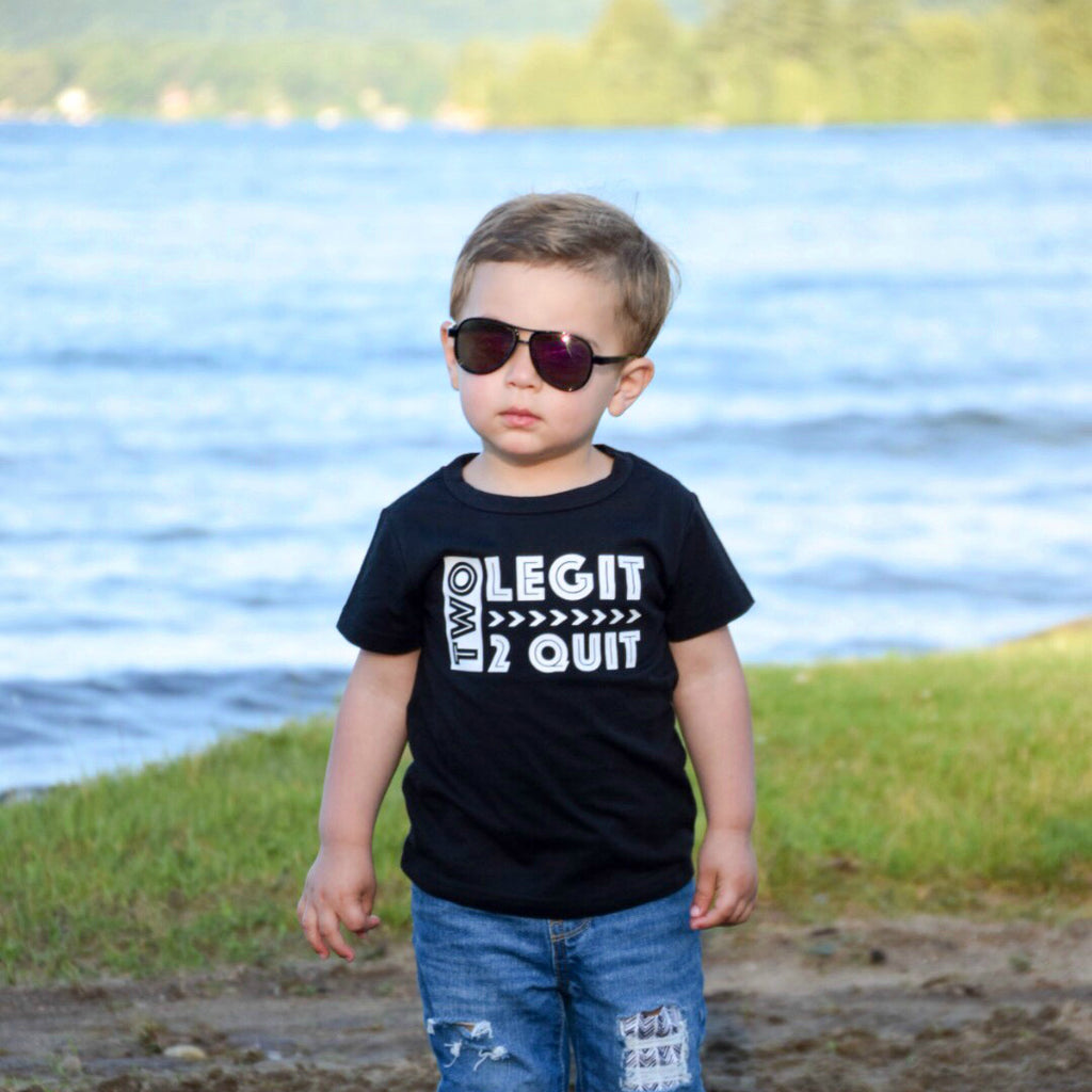 Little boy in front of lake wearing sun glasses and black shirt that says Two Legit 2 Quit in white