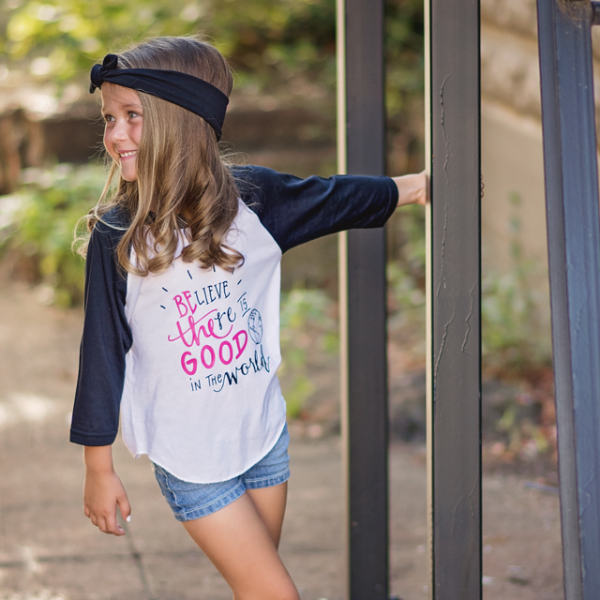 Little girl wearing black sleeve raglan that says Believe There Is Good In The World in pink and black