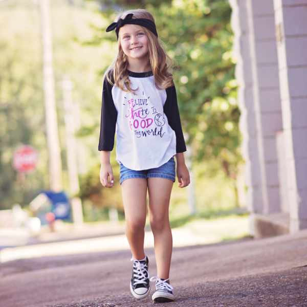 Little girl wearing black sleeve baseball tee that says Believe there is good in the world in black and pink